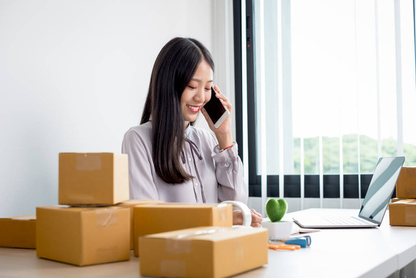 Fiatal ázsiai nő üzleti tulajdonos sok csomag dobozok az asztalon boldog online értékesítési munkát, használja az okostelefon, kap egy megrendelést az ügyfelek, jegyzetek, és intézkedjen a szállítás postai úton. - Fotó, kép