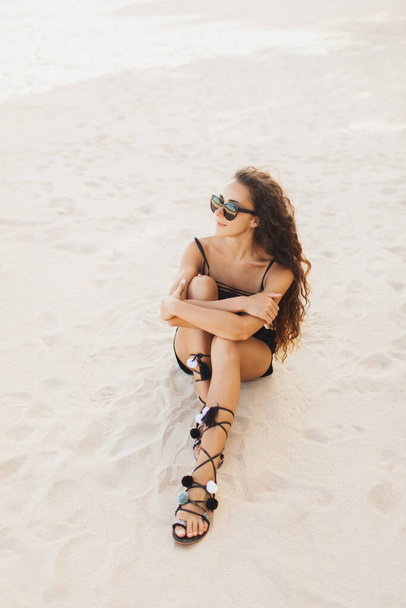Νεαρή μελαχρινή γυναίκα με μακριά μαλλιά σε γυαλιά ηλίου και μαύρο μαγιό που χαλαρώνει στην παραλία και κάθεται σε λευκή άμμο το ηλιοβασίλεμα. - Φωτογραφία, εικόνα
