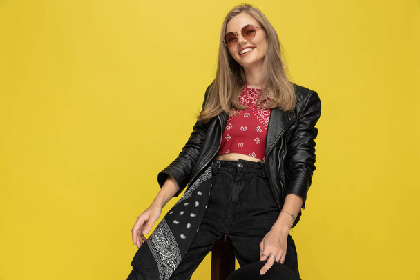 Happy fashion model wearing sunglasses and smiling while sitting on a chair on yellow studio background - Photo, Image