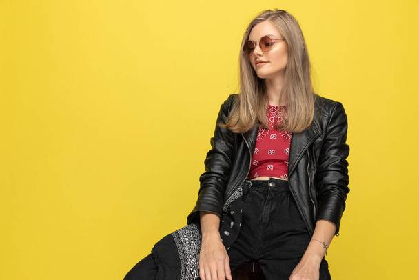 Positive fashion model wearing sunglasses and smiling, looking away while sitting on a chair on yellow studio background - Фото, зображення