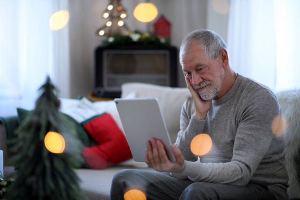 Sad and lonely senior man with tablet sitting at the table indoors at Christmas, solitude concept. - Foto, Imagen
