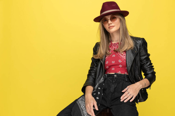 Bossy fashion model wearing hat and sunglasses while sitting on a chair on yellow studio background - Photo, Image