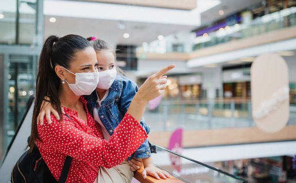 Mother and daughter with face mask standing indoors in shopping center, coronavirus concept. - Fotografie, Obrázek