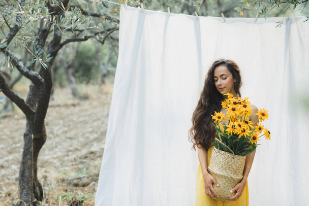 Portrait of young brunette woman on background of white hanging textile cloth in garden. Holding straw basket with yellow flowers, summer linen dress. - Photo, Image