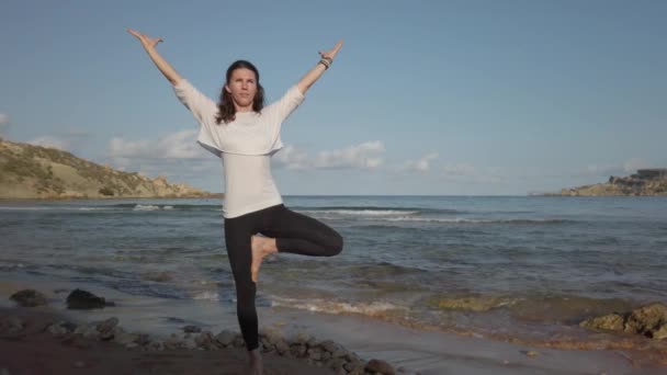 Frau steht auf einem Bein, während sie Yoga an einem Sandstrand praktiziert - Filmmaterial, Video