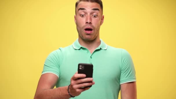 junger, lässiger Mann schaut auf sein Telefon, ist schockiert, macht Welpenaugen, schlägt sein Gesicht auf gelbem Hintergrund - Filmmaterial, Video
