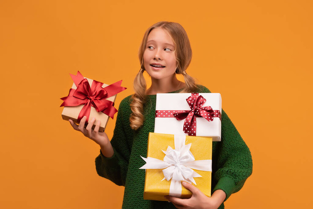 Imagen de la encantadora chica rubia de 12-14 años de edad en suéter verde cálido sosteniendo una gran cantidad de cajas de regalo. Fotografía de estudio, fondo amarillo, aislado. Año Nuevo Día de la Mujer Cumpleaños Concepto de vacaciones - Foto, Imagen