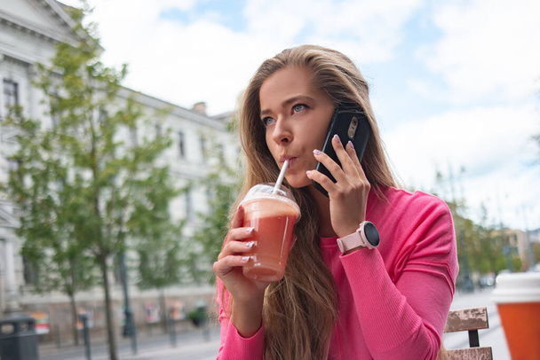 Woman using a straw to drink grapefruit juice outdoor in a city street while talking on the phone - Foto, imagen