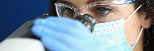 Woman in protective mask and gloves looks through microscope. - 写真・画像