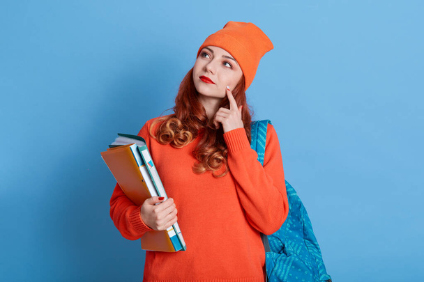 Portrait of pensive Caucasian girl with thoughtful expression, concentrated on important task, looks aside with finger on cheek, dressed in orange jumper and cap, goes to college. - Foto, Bild