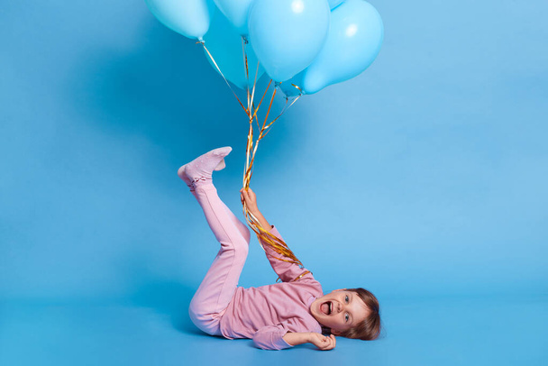 Little toddler girl having fun while lying on floor with raised legs, looks at camera and yelling happily, female kid plays with balloon against blue wall, child in pink casual attire. - Photo, image
