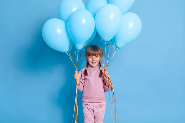 Funny attractive female kid goes on birthday party, carries bunches helium balloons, wears pink jumper and pants, child with pigtails smiles broadly, isolated on blue wall. - Photo, image