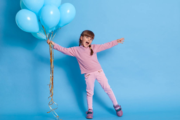 Fille avec un tas de ballons d'hélium écartant les mains et criant quelque chose de heureux, portant pull rose décontracté, pantalon et baskets sur fond bleu, enfant mignon à la fête. - Photo, image