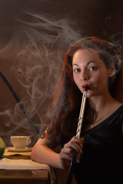 Jovem mulher bonita em vestido preto fuma um narguilé ou shisha na boate ou bar fumaça. Fumaça e vapor no ar. - Foto, Imagem