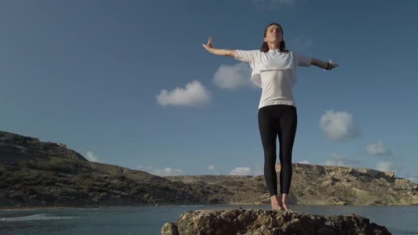 Jonge blanke vrouw doet yoga ademhaling en stretching oefeningen op rots op het strand - Video