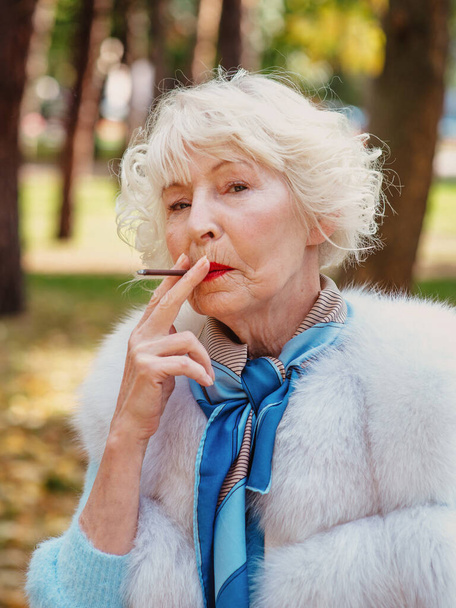 triste mujer mayor de moda con el pelo gris en abrigo de piel fumar cigarrillos al aire libre. Estilo de vida poco saludable, edad, vejez, adicción, mal concepto de hábito. - Foto, Imagen
