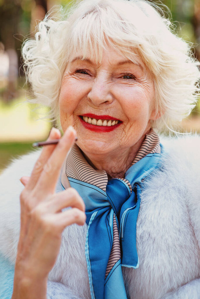 glimlachende senior stijlvolle modieuze vrouw met grijs haar in bontjas buiten roken sigaret. Ongezonde levensstijl, leeftijd, positieve vibes, ouderdom, verslaving, slechte gewoonte concept. - Foto, afbeelding