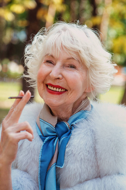 smiling senior elegant stylish fashionable woman with grey hair in fur coat outdoor smoking cigarette. Unhealthy lifestyle, age, positive vibes, oldness, addiction, bad habit concept. - Photo, Image