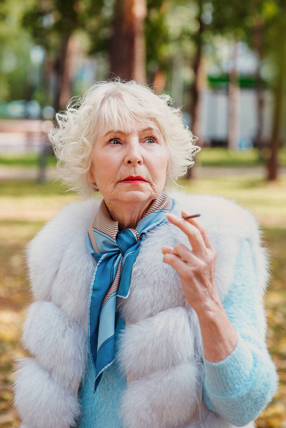 θλιβερή μοντέρνα ηλικιωμένη γυναίκα με γκρίζα μαλλιά σε γούνα παλτό υπαίθριο κάπνισμα τσιγάρο. Ανθυγιεινός τρόπος ζωής, ηλικία, ηλικία, εθισμός, έννοια της κακής συνήθειας. - Φωτογραφία, εικόνα