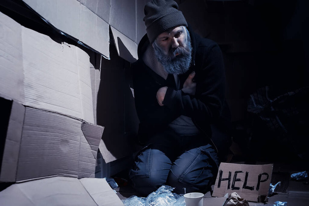 Бездомный бородатый сидит на коробках на улице и просит о помощи. Бездомный просит деньги на еду и ночлег. - Фото, изображение