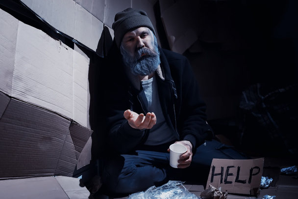 Ein bärtiger Obdachloser sitzt auf Kisten auf der Straße und bittet um Hilfe. Ein Obdachloser bittet um Geld für Essen und Übernachtung. - Foto, Bild
