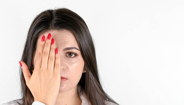 Young brunette woman   covering one eye with her hand and looking at camera. Front portrait against gray background with copy space - Foto, Imagen