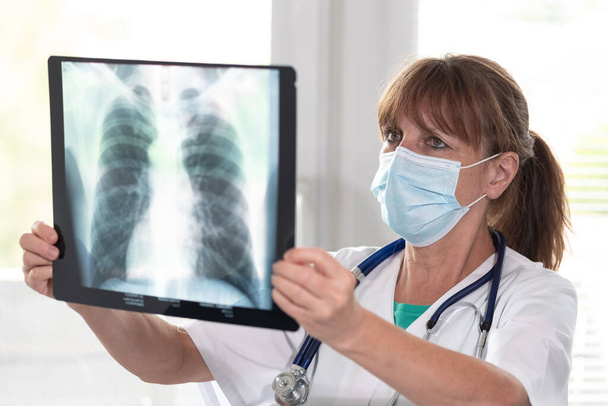 Женщина-врач носит медицинскую маску для лица и изучает рентген легких во время вспышки коронавируса - Фото, изображение