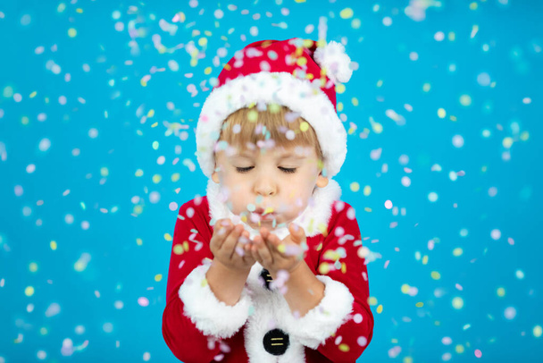 Happy child wearing Santa Claus costume. Kid blowing confetti against blue background. Christmas holiday concept - Foto, Imagem