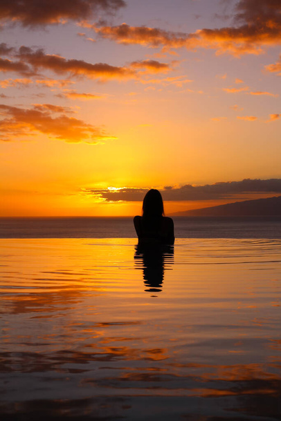 una chica está de pie con su espalda a la piscina, sobre el fondo de una puesta de sol naranja y el océano. - Foto, Imagen