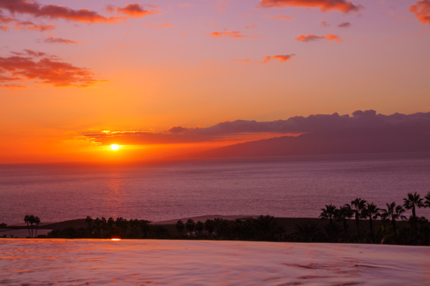 Blick von oben auf den Pool, einen Uferstreifen mit Palmen, das Meer und die Insel bei Sonnenuntergang - Foto, Bild