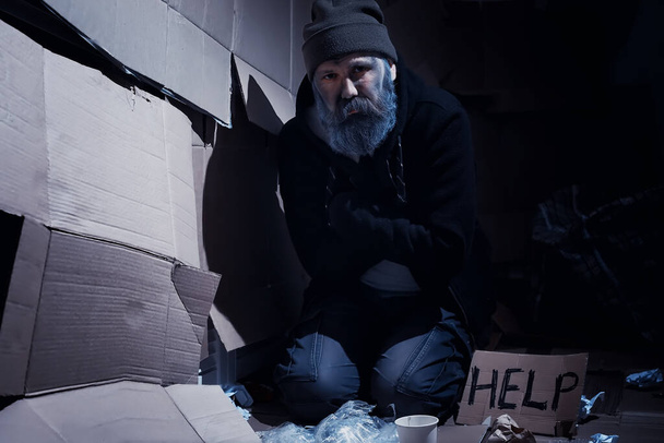 Bezdomny brodaty mężczyzna siedzi na pudłach na ulicy i prosi o pomoc. Potrzebujesz bezdomnego prosi o pieniądze na jedzenie i nocleg. - Zdjęcie, obraz