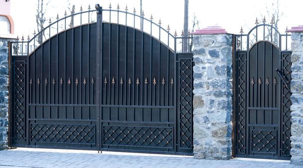 Zwarte metalen poorten en deuren met een gesmeed patroon en een deel van een stenen hek. - Foto, afbeelding