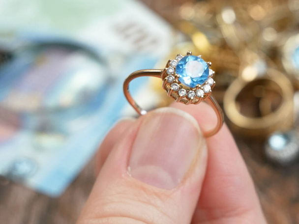 χέρι παίρνει δαχτυλίδι με μεγάλη πέτρα topaz και πολλά διαμάντια, θραύσματα κοσμημάτων από χρυσό και ασήμι και χρήματα, ενεχυροδανειστήριο έννοια, closeup - Φωτογραφία, εικόνα
