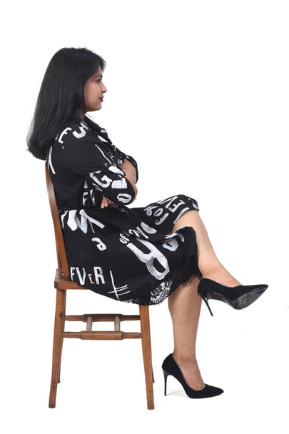 Beyaz arka planda sandalyede oturan, kolları ve bacakları çaprazlanmış, yüksek topuklu elbiseli Latin kadının yan görüntüsü. - Fotoğraf, Görsel