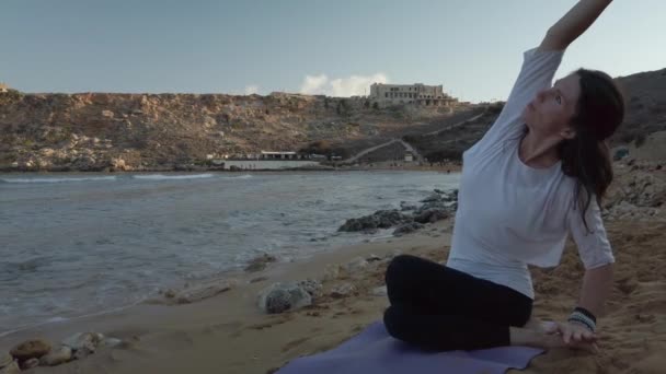 Женщина за 40 практикует йогу на песчаном пляже рано утром - Кадры, видео