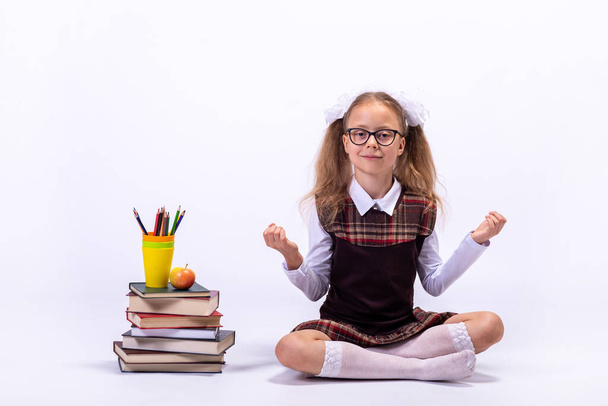A little girl in a school uniform and glasses sits on a white background next to a pile of books. School concept. - Foto, Imagen
