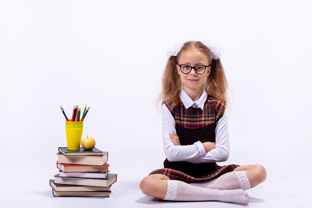 A little girl in a school uniform and glasses sits on a white background next to a pile of books. School concept. - Foto, Imagem
