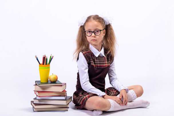 A little girl in a school uniform and glasses sits on a white background next to a pile of books. School concept. - Zdjęcie, obraz