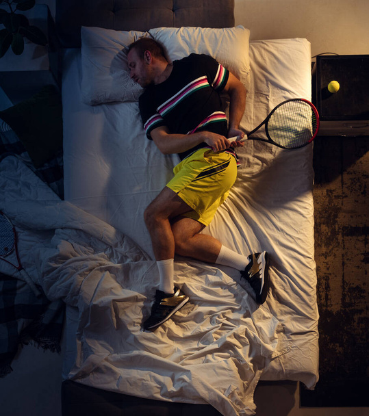 Κάτοψη του νεαρού επαγγελματία τενίστα που κοιμάται στην κρεβατοκάμαρά του με αθλητικά ρούχα και ρακέτα - Φωτογραφία, εικόνα