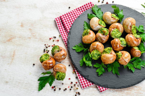 Baked snails with garlic butter and fresh herbs on a black plate on a white wooden background. Top view. Free space for your text. - Fotoğraf, Görsel
