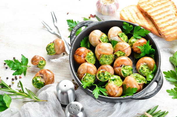 Baked snails with garlic butter and fresh herbs on a black plate on a white wooden background. Top view. Free space for your text. - 写真・画像