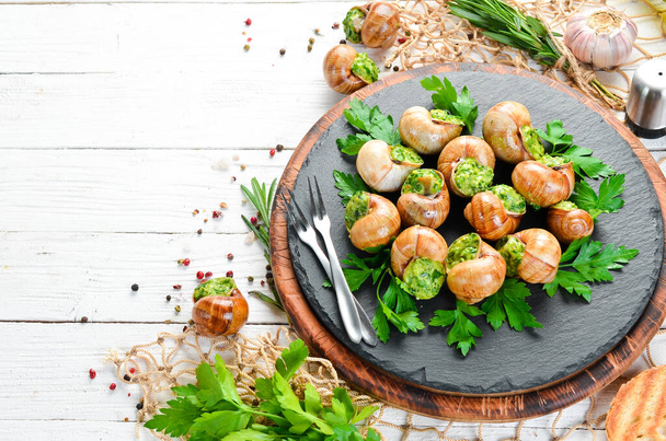 Baked snails with garlic butter and fresh herbs on a black plate on a white wooden background. Top view. Free space for your text. - Foto, Bild