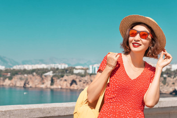 Uma mulher atraente em um chapéu de palha e vestido vermelho relaxa e admira as vistas da estância marítima mediterrânea e Riviera. Conceito de viagem e férias - Foto, Imagem
