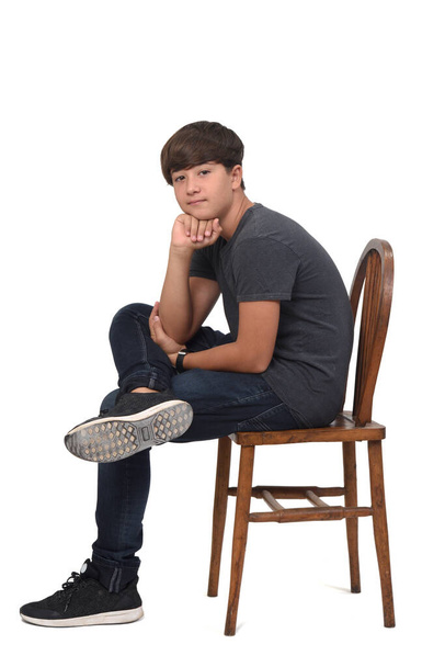 nastolatek chłopiec siedzi na krześle z białym tle, patrząc w aparat fotograficzny i ręka na brodzie - Zdjęcie, obraz