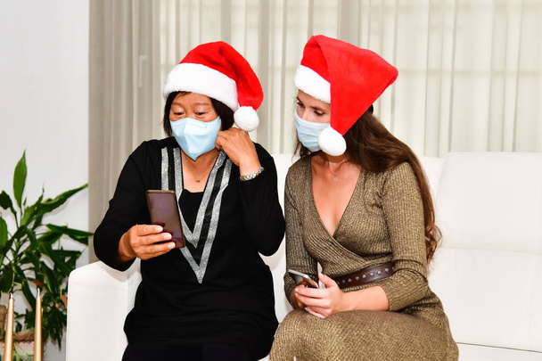 Kobiety noszące czapki Mikołaja i maski na twarz patrzące na telefon podczas relaksu na kanapie. Święta Bożego Narodzenia i koncepcja bezpieczeństwa. - Zdjęcie, obraz