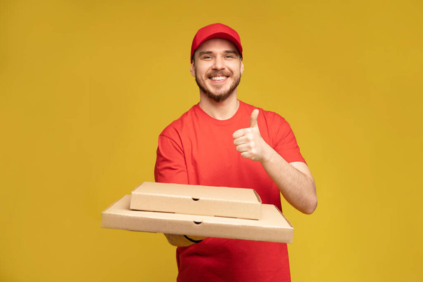 Happy man from delivery service in red t-shirt and cap giving food order and holding pizza box isolated over yellow - Foto, Imagem