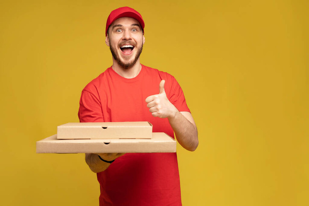 Happy man from delivery service in red t-shirt and cap giving food order and holding pizza box isolated over yellow - Photo, Image