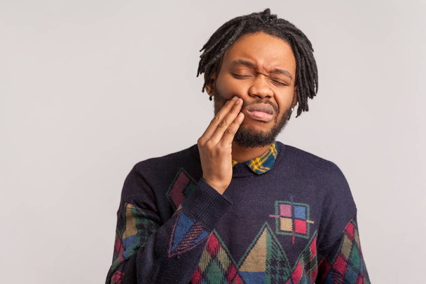 Frustrated bearded african man with dreadlocks touching cheek and wincing in pain feeling terrible tooth ache, gingivitis, periodontal disease. Indoor studio shot isolated on gray background - Foto, imagen