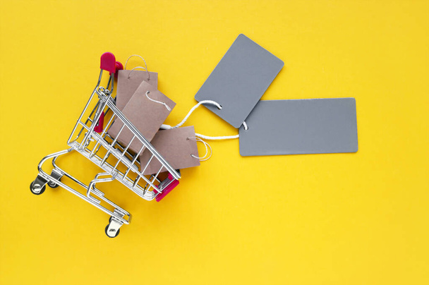 Küçük el çantaları ve sarı arka planda boş gri etiketleri olan mini alışveriş arabası. Mağazalarda ve internette alışveriş kavramı. - Fotoğraf, Görsel
