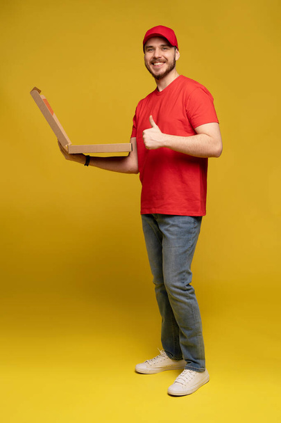Onnellinen mies jakelupalvelusta punaisessa t-paidassa ja lakissa, joka antaa ruokatilauksen ja pitää pizzalaatikkoa eristettynä keltaisen päälle - Valokuva, kuva